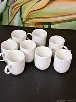 Group photo showing 8 white textured ceramic mugs arranged on a black surface in front of a colorful background.