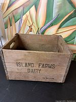 Front and side angled view of vintage wooden dairy box with 'Island Farms Dairy 1/2 Gallon' stenciled on side. Shows rectangular shape, cut-out handles, and metal corner reinforcements.