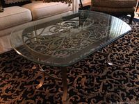 Front angle view of rectangular glass top coffee table on patterned carpet, showing metal frame and scrollwork design.