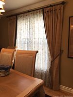 View of 2 beige drapery panels hanging on black curtain rod, pulled back with tassel tiebacks, sheer curtain behind not included.