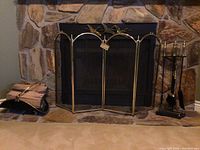 Wide view of the fireplace with brass bird screen at center, log holder on left holding firewood, and brass tool set on the right.