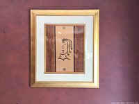 Full view of framed abstract art print with beige panel, brown side panels, black abstract line face drawing, white mat, and gold frame