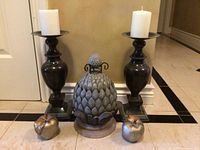 Full view of the candle holders, artichoke sculpture, and apples arranged on floor against wall.