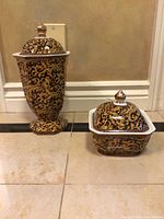Pair of decorative ceramic jars with lids, one tall and one square, both with intricate black and gold floral designs, placed on tile floor.