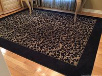 Full view of the black bordered rug with beige floral scroll pattern, laid under dining furniture on hardwood floor.