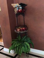 Full view of black metal stand with two wire baskets projected on painted brown wall with decorative curly metalwork and a metal scrolled base.