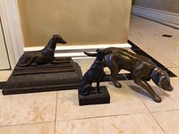 Photo showing three bronze dog statues of varying sizes and poses placed on a tile floor against a wall.