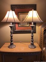 Pair of ornate metal base table lamps with white fabric shades placed on a light wood cabinet.
