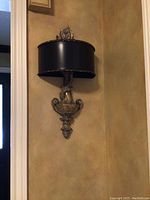 Full view of one wall sconce mounted on wall showing decorative urn-shaped base and black shade