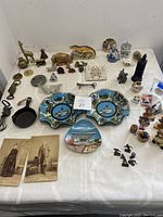 Overview of the 52 vintage small collectibles spread on a table showing various brass, ceramic, and metal items including commemorative plates, photographs, figurines, and small kitchenware.
