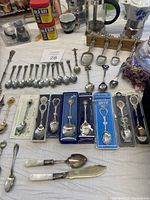 Collection of collectible spoons, including Canadian-themed teaspoons and measuring spoons mounted on wooden rack, along with kitchen items including a moka pot, salt and pepper shakers, and Old Bay seasoning cans.