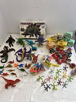 Wide view showing assorted dinosaur figurines, dinosaur and lizard party favor toys, Flintstone family figurines and cars, unopened Stegosaurus model box, and other pieces collected on a white surface.