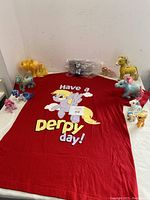 Red T-shirt laid flat with 'Have a Derpy Day!' graphic featuring a winged pony character, surrounded by eight assorted My Little Pony figurines on a white surface.