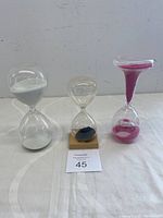 Three hourglass timers displayed together, showing variety in sand colors and sizes, including the magnetic hourglass on wooden base.
