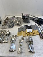 Photo showing assorted vintage forks, spoons, knives with cream-colored handles, some silver plated items in containers, and steak knives with black and wooden handles.