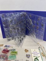 Open blue binder showing clear pockets filled with a variety of charms, plus several small bags of colorful beads and stones placed in front