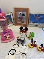 Photo showing Minnie Mouse vacuum cleaner, framed pictures of Mickey Mouse and Dumbo, two View-Master viewers with multiple vintage Disney slide reels, and two Mickey Mouse fridge magnets on a table.