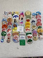 Full view of a collection of 62 assorted vintage advertising and souvenir buttons arranged in rows on a light fabric surface. Buttons vary in color, size, and theme with legible text and graphics focused on Canadian organizations and campaigns.
