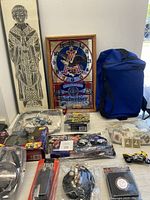 Photo showing entire lot including grave rubbing art in frame, Budweiser mirror clock, blue sports bag, motorcycle collectibles in packaging, Canadian stamps, coins, and various small items on table.