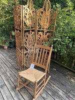 Photo showing full view of rattan folding room divider and wooden rocking chair placed outdoors on deck with greenery background.