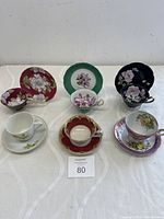 All six matched pairs of vintage hand painted teacup and saucer sets displayed together on a flat surface.