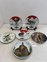 Six collector plates displayed on a table, including three Wolpertinger themed plates, one with a bird, one with a deer, and one with a Bavarian building.