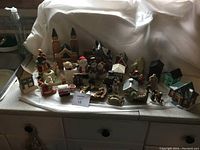 Wide shot showing the full collection of ceramic village buildings and figurines arranged on a surface with a white backdrop, highlighting the variety and scale of the items.