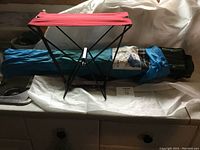 Two camping chairs with fabric seats and foldable black metal frames, accompanied by their blue and green carrying bags.