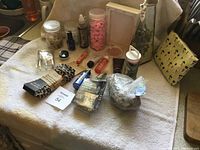 Photo showing an assortment of craft materials including jars with fillers, LED string lights, small tubes and bottles, and fabric pieces on a towel.