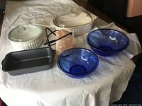 Photo showing two white ceramic casserole dishes with lids, two blue glass mixing bowls, small gray loaf pan, and pink salt lamp with cord on white cloth.