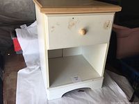 Full view of the night stand illustrating the natural wood top, white painted body, drawer with knob, open shelf, and sticker marks on drawer front.