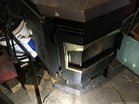 Black pellet stove with metallic front door and glass window, positioned on floor in dark surroundings.