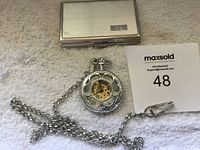 Silver pocket watch on chain next to silver business card case on white fabric background.