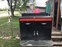 Front view of the black and orange Black and Decker tool chest showing two top drawers with handles and a double-door cabinet below, placed on a glass table outdoors.