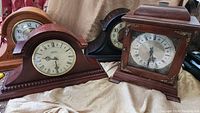 Four vintage mantel clocks displayed on a cloth, showcasing different wood finishes and shapes.