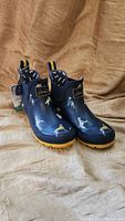 Front and side view of Joules navy blue rain boots with dog pattern and yellow soles on beige fabric background.