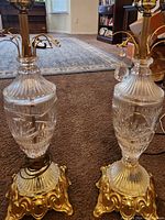 Pair of vintage Accurate Casting lamps with clear cut glass urn bases, gold-tone metal ornate pedestal stands, and hanging crystal prisms, sitting on carpet indoors.