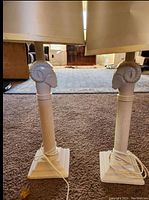 Photo of two cream-colored vintage table lamps with detailed rams head design on lamp posts and beige fabric lampshades standing on carpeted floor.