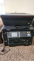 Front and top view of Epson WorkForce WF-3620 printer showing scanner lid, control panel with LCD screen and keypad, resting on carpeted floor.