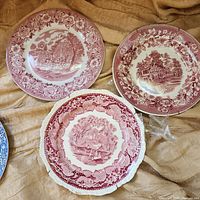 Top-down view of 3 vintage red transferware China dinner plates on fabric background, showing distinct scenic patterns and floral borders.