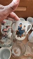 Hand holding clear glass tumbler with pink Ice Age Sid the Sloth character print next to Norman Rockwell ceramic mug with seated elderly man and child illustration