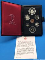 Open red Royal Canadian Mint presentation booklet showing eight proof coins arranged in black foam including two dollar coins and other denominations from penny to half dollar along with a certificate on a blue background.