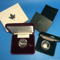Two Canadian dollar coins in original presentation boxes, one white outer box with minor staining, maroon and dark green velvet inner boxes, coins in clear capsules.