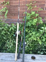 Three fishing rods leaning against a brick wall and green foliage on wooden surface showing cork and synthetic handles and reels attached