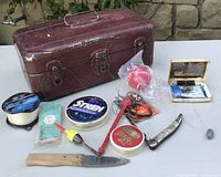 Photo showing worn red metal tackle box with assorted fishing gear spread out around it.