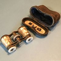 Vintage opera glasses with brass and mother of pearl inlay shown alongside open brown leather hard shell carry case with fabric interior.