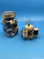 Full view of the needle and thread carousel alongside the Mason jar full of assorted buttons on a blue background.