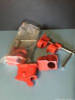 Two red cast metal pipe clamp heads with threaded screws and T-handles, new in plastic bags, placed on table