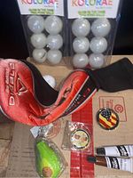 Two packages of glow in the dark ping pong balls, a red and black golf club cover, green squeegee keychain in packaging, and two acrylic paint tubes with two round pins on cardboard surface.