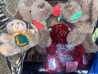 Four Valentine-themed stuffed animals including two brown dogs holding red roses, one red dog with 'Love' and heart decorations, and one cream bear with a green and orange letter 'L' on its chest. Includes a red decorative pouch.